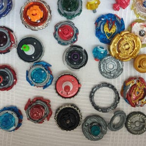 Op de afbeelding: Een verzameling kleurrijke tolletjes in verschillende ontwerpen en maten. De tolletjes hebben een mix van rode, blauwe, gouden en zilveren accenten. Sommige hebben ingewikkelde patronen en gelaagde componenten, andere eenvoudigere ontwerpen.