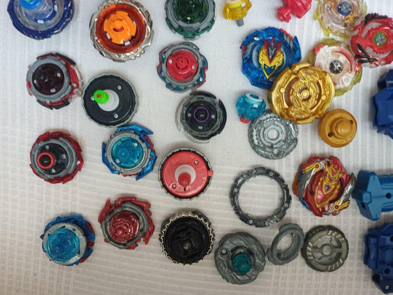 Op de afbeelding: Een verzameling kleurrijke tolletjes in verschillende ontwerpen en maten. De tolletjes hebben een mix van rode, blauwe, gouden en zilveren accenten. Sommige hebben ingewikkelde patronen en gelaagde componenten, andere eenvoudigere ontwerpen.