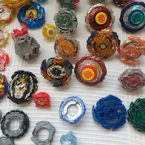 Op de afbeelding: Een verzameling kleurrijke tolletjes in verschillende ontwerpen en maten. De tolletjes hebben een mix van rode, blauwe, groene, gele en transparante plastic componenten. Sommige hebben ingewikkelde patronen en details, andere zijn eenvoudiger van ontwerp.