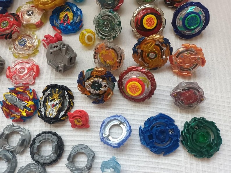 Op de afbeelding: Een verzameling kleurrijke tolletjes in verschillende ontwerpen en maten. De tolletjes hebben een mix van rode, blauwe, groene, gele en transparante plastic componenten. Sommige hebben ingewikkelde patronen en details, andere zijn eenvoudiger van ontwerp.