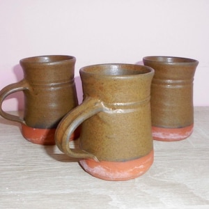 Op de afbeelding: Drie keramische mokken met een tweekleurig ontwerp, voorzien van een donkerbruin geglazuurd bovendeel en een ongeglazuurde terracotta-kleurige basis. Elke mok heeft een gebogen handvat en een licht uitlopende rand in cilindrische vorm.