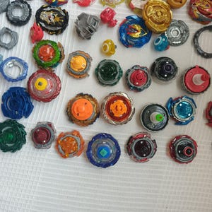 Op de afbeelding: Een verzameling kleurrijke tolletjes in verschillende ontwerpen en maten. De tolletjes hebben een mix van kleuren, waaronder blauw, rood, groen, oranje en goud. Sommige hebben ingewikkelde patronen, andere zijn eenvoudiger.