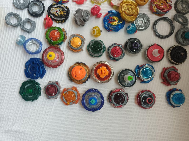 Op de afbeelding: Een verzameling kleurrijke tolletjes in verschillende ontwerpen en maten. De tolletjes hebben een mix van kleuren, waaronder blauw, rood, groen, oranje en goud. Sommige hebben ingewikkelde patronen, andere zijn eenvoudiger.