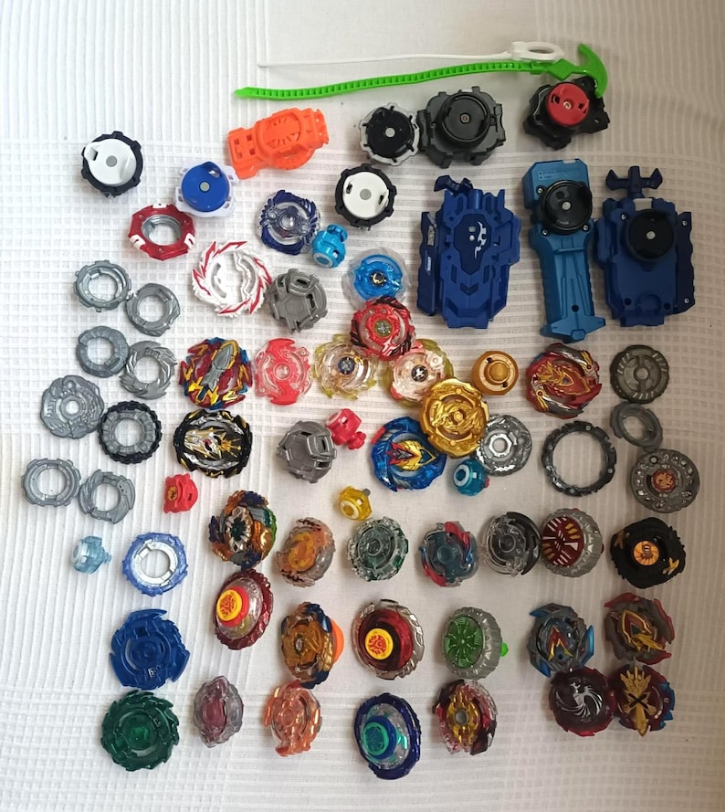 Op de afbeelding: Een verzameling kleurrijke tol speelgoed, inclusief lanceerders en diverse componenten. De toppen hebben een verscheidenheid aan ontwerpen en kleuren, zoals rood, blauw, goud en groen. De afbeelding toont een verscheidenheid aan maten en vormen.