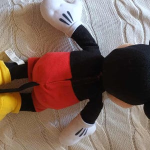 May include: A plush Mickey Mouse doll, lying on its back, features a black head with large ears, a red pair of shorts, yellow shoes, and white gloves. The doll is made of soft fabric and is a classic children's toy.