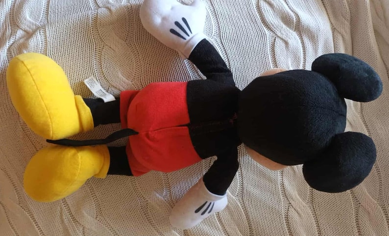 May include: A plush Mickey Mouse doll, lying on its back, features a black head with large ears, a red pair of shorts, yellow shoes, and white gloves. The doll is made of soft fabric and is a classic children's toy.