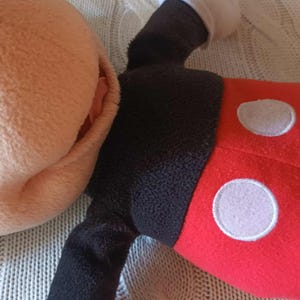 May include: A plush Mickey Mouse toy, featuring a tan face and ears, black eyes, and a black body. The toy's red shorts have two white circles. The toy is lying on a textured, light-colored surface.
