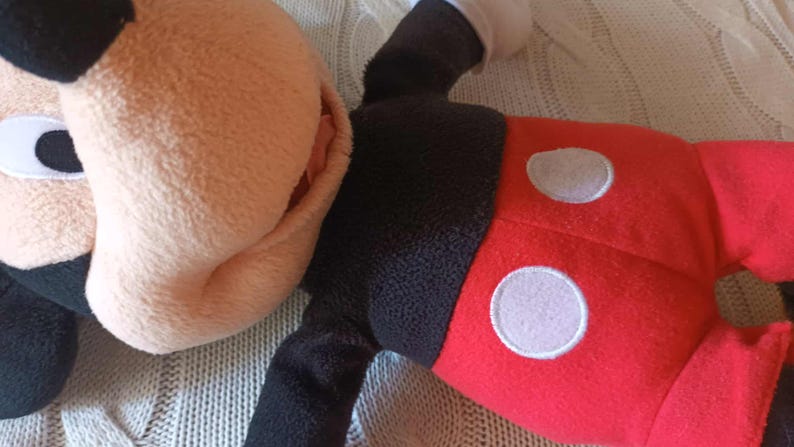 May include: A plush Mickey Mouse toy, featuring a tan face and ears, black eyes, and a black body. The toy's red shorts have two white circles. The toy is lying on a textured, light-colored surface.