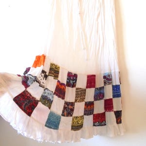 May include: White maxi skirt with a patchwork hem featuring colorful squares of patterned fabric. The skirt has a ruffled hemline and a drawstring waist. The skirt is made of a lightweight fabric.