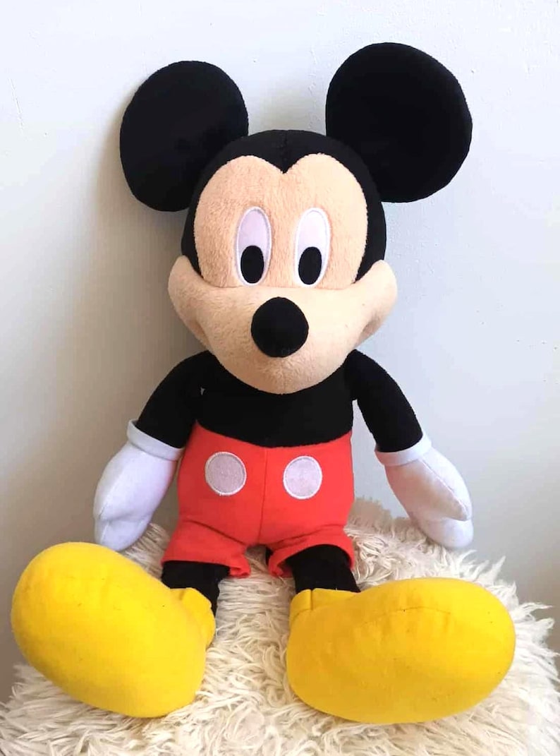 May include: A plush Mickey Mouse doll, featuring black ears, a black body, red shorts with white circles, and large yellow shoes. The doll has a tan face, black eyes, and white gloves. It is sitting on a fluffy, white surface.