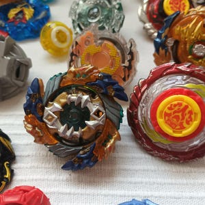 Op de afbeelding: Een verzameling kleurrijke tolletjes in verschillende ontwerpen. De tolletjes hebben een mix van rode, blauwe, gouden en zilveren elementen. Sommige hebben ingewikkelde gelaagde ontwerpen, terwijl andere eenvoudigere, effen kleurenschema's hebben. De tolletjes zijn gerangschikt op een wit oppervlak.