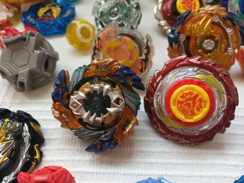 Op de afbeelding: Een verzameling kleurrijke tolletjes in verschillende ontwerpen. De tolletjes hebben een mix van rode, blauwe, gouden en zilveren elementen. Sommige hebben ingewikkelde gelaagde ontwerpen, terwijl andere eenvoudigere, effen kleurenschema's hebben. De tolletjes zijn gerangschikt op een wit oppervlak.