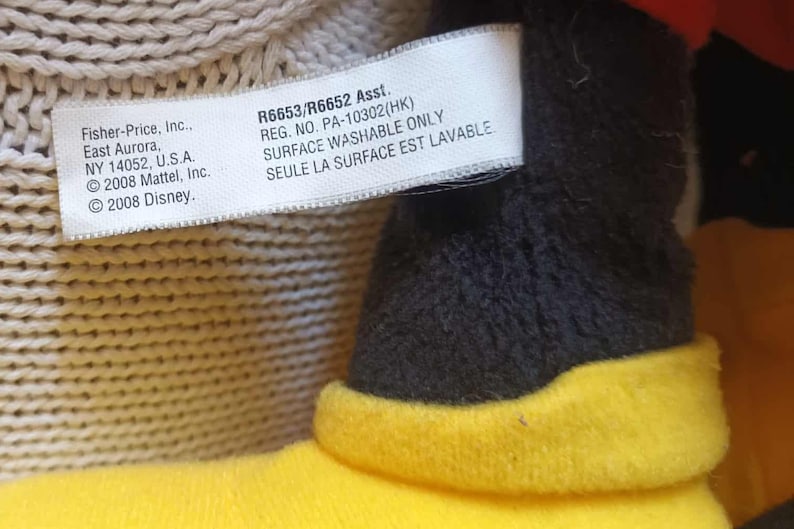 May include: Close-up of a plush toy's tag with text from Fisher-Price, Inc., including the address in East Aurora, NY, and copyright information for Mattel and Disney from 2008. The tag also includes the text "SURFACE WASHABLE ONLY".