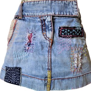 May include: Light blue denim skirt with visible patches and decorative stitching. Patches are in various colors, including black, red, and purple. The skirt has a classic five-pocket design and a button and zipper closure.
