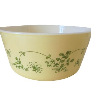 Op de afbeelding: Een vintage gele mengkom met een witte binnenkant en twee handvatten. De kom heeft een groen bloemenpatroon aan de buitenkant. Dit keukengerei is ideaal voor bakken en het bereiden van voedsel.