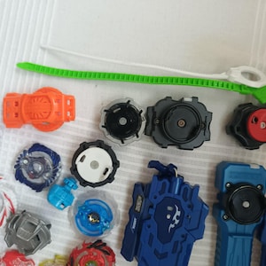 Op de afbeelding: Een verzameling kleurrijke tol speelgoed en lanceerders. De toppen hebben verschillende ontwerpen en kleuren, waaronder blauw, rood, wit en zilver. Lancers zijn oranje en blauw, met een groen en wit trekkoord.