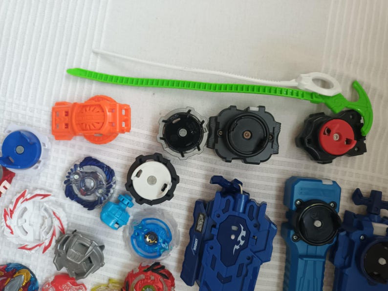 Op de afbeelding: Een verzameling kleurrijke tol speelgoed en lanceerders. De toppen hebben verschillende ontwerpen en kleuren, waaronder blauw, rood, wit en zilver. Lancers zijn oranje en blauw, met een groen en wit trekkoord.