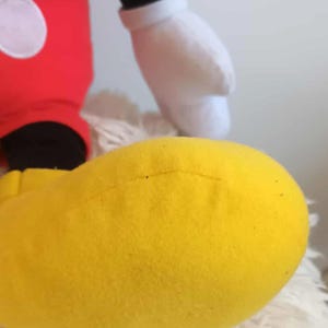 May include: A plush toy featuring a large yellow foot, red pants with a white circle, a black leg, and a white glove. The toy is a recognizable character from a popular animated series.