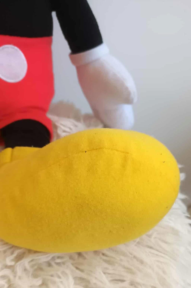 May include: A plush toy featuring a large yellow foot, red pants with a white circle, a black leg, and a white glove. The toy is a recognizable character from a popular animated series.