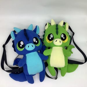 May include: Two plush dragon backpacks. One is blue with a light blue belly, and the other is green with a yellow belly. Both have large, round eyes with black pupils and green accents. They have black adjustable straps.