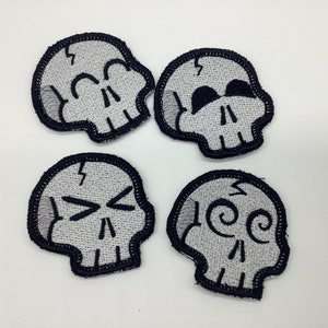 May include: Four embroidered iron-on patches featuring different skull designs. Each skull is white with black outlines and features different facial expressions.