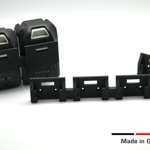 Könnte beinhalten: Schwarze Akku-Werkzeug-Akkupacks und Montagehalterungen. Die Akkupacks haben ein weißes und schwarzes Design mit rechteckiger Form. Die Montagehalterungen sind schwarz und haben eine rechteckige Form mit Löchern. Der Text "Made in Germany" ist sichtbar.