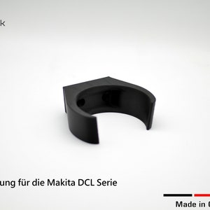 Könnte beinhalten: Eine schwarze Wandhalterung aus Kunststoff für ein Makita DCL-Serienwerkzeug. Die Halterung ist C-förmig und hat zwei kleine Löcher auf der Rückseite. Der Text "1x Halterung für die Makita DCL Serie" ist unter der Halterung aufgedruckt. Der Text "Made in Germany" ist unter dem Text in Schwarz, Rot, Gelb und Schwarz aufgedruckt.