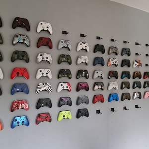 Op de afbeelding: Een wanddisplay met meer dan 50 kleurrijke Xbox-controllers die aan zwarte haken zijn bevestigd. De controllers zijn gerangschikt in rijen en kolommen, met een verscheidenheid aan kleuren en ontwerpen.