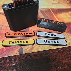 May include: A black plastic box with a label that says "UKIEBOX" and a set of orange and black tokens. There are four labels that say "ACTIVATION", "CREW", "TRIGGER", and "UNTAP".