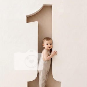 May include: A young child with light brown hair stands inside a large, three-dimensional cutout of the number one. The child is wearing a light beige sleeveless romper. The structure is off-white with a smooth, matte finish and deep interior.