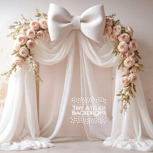 May include: A white fabric backdrop with a large white bow at the top and a garland of blush pink roses and greenery on each side. The backdrop is draped in a way that creates a soft, romantic archway. The text "Tiny Atelier Backdrops" is printed in white on the backdrop.