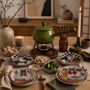 May include: A green fondue pot with a wooden handle sits on a black metal stand over a flame. The table is set with plates featuring cat illustrations, a bowl of mushrooms, a bowl of bread cubes, a candle, and a vase with branches.