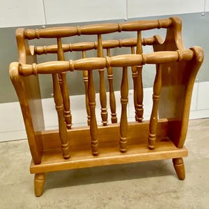 May include: A wooden magazine rack with a light brown finish. The rack features a slatted design with vertical spindles and horizontal bars. The sides are solid and curved, and the rack has four short legs.