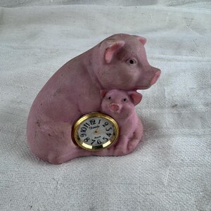 Może przedstawiać: Matowa, różowa ceramiczna figurka przedstawiająca dużą świnię stojącą nad prosiakiem. Okrągły analogowy zegar kwarcowy ze złotą metalową ramką i białą tarczą z czarnymi cyframi oraz nazwą marki Xonox jest wbudowany w bok figurki świni.