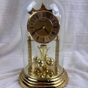 May include: A gold-toned clock with a glass dome. The clock face has Roman numerals and the brand name "Junghans". The base and decorative elements are gold, and the clock features a pendulum with multiple gold spheres. The clock is a decorative item.