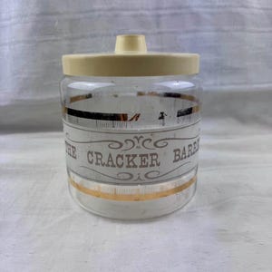 Puede incluir: Un tarro de cristal transparente con tapa de plástico color crema. El tarro presenta una banda blanca central con el texto THE CRACKER BARREL impreso en marrón, decorado con volutas ornamentales y finas rayas horizontales doradas.