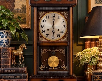 Op de afbeelding: Een donkerbruine houten schoorsteenklok met een rechthoekig frame, een sierlijke bovenkant en een ronde wijzerplaat met zwarte Arabische cijfers. Het onderste gedeelte heeft een glazen paneel met een ronde koperen slinger en gouden details.