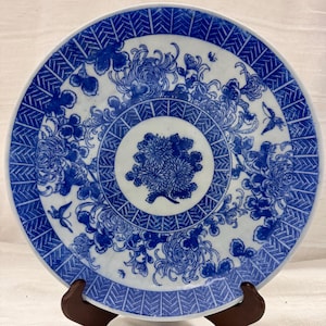 Puede incluir: Un plato redondo de cerámica blanca con un diseño floral azul. El plato presenta un motivo floral central rodeado de intrincados patrones y elementos decorativos. El borde tiene un borde geométrico. El plato se exhibe sobre un soporte marrón.