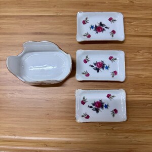 Op de afbeelding: Een set van vier witte porseleinen schalen met een bloemmotief. De set bevat drie rechthoekige schalen en een uniek gevormde schaal. Elke schaal heeft een gouden rand en een patroon van roze rozen en blauwe accenten.