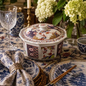 May include: A white porcelain lidded bowl with an octagonal shape, decorated with intricate blue, red, and gold floral and geometric patterns. The bowl sits on a patterned tablecloth alongside a crystal glass, a napkin ring, and plates.