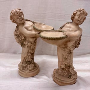 Puede incluir: Dos estatuas de querubines beige sosteniendo cuencos redondos y poco profundos. Los querubines están adornados con vides y hojas de uva. Los cuencos tienen un borde en forma de cuerda. Las estatuas están sobre una base redonda.