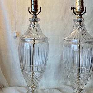 May include: Two clear glass table lamps with a ribbed design and diamond pattern. The lamps have brass-colored hardware and are topped with light bulbs. The lamps are on a white surface.