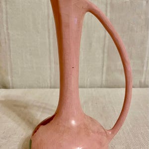 Puede incluir: Un jarrón de cerámica rosa vintage con un asa única. El jarrón tiene una base bulbosa y un cuello alto y delgado que se curva en un asa. La superficie tiene un esmalte craquelado. El jarrón es de color rosa claro.