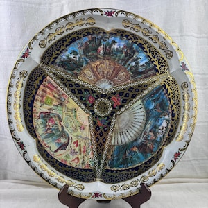 Puede incluir: Una bandeja decorativa redonda con tres secciones, con obras de arte con temática de abanicos. La bandeja tiene una base blanca con detalles dorados y azul marino, y patrones florales y ornamentados. La obra de arte representa escenas con personas y abanicos.