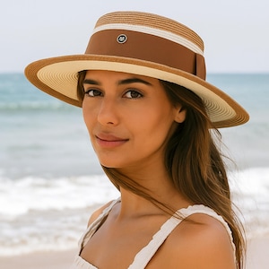 May include: A straw hat with a wide brim and a brown band. The hat has a white stripe and a small emblem on the band. The hat is worn by a person on a beach with the ocean in the background.