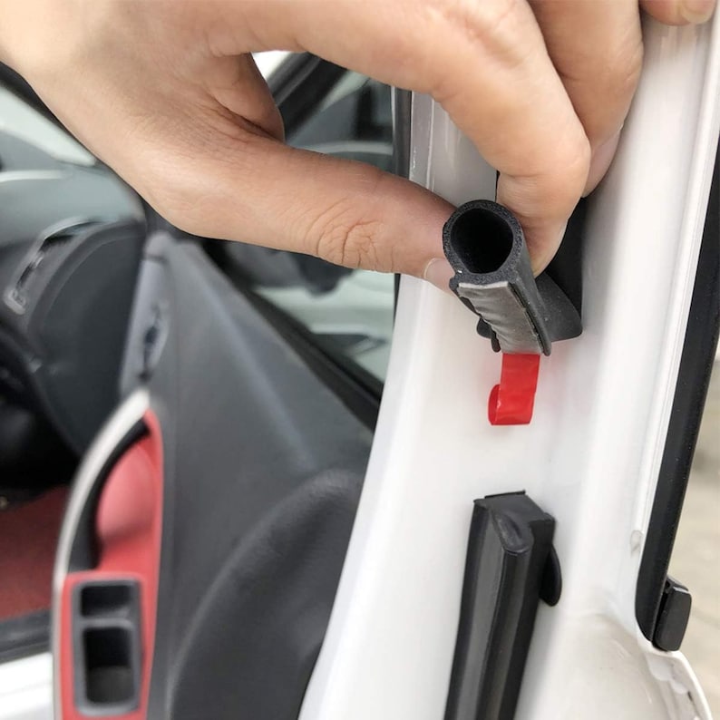 May include: A close-up of a black rubber seal being installed in a car door. The seal has a red tab and is being held in place by a hand.