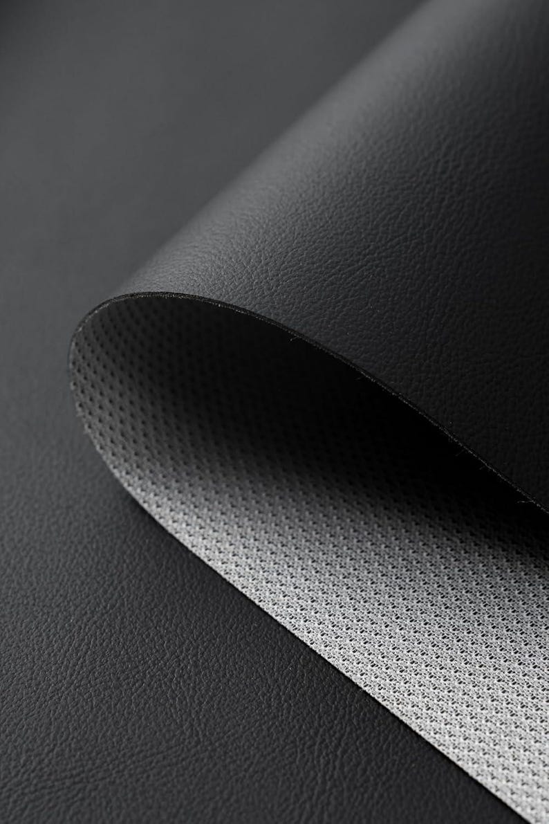 May include: Close-up of a black leather material, partially rolled to reveal a gray, textured underside. The leather has a subtle grain, and the overall effect is one of depth and texture. The image highlights the contrast between the smooth exterior and the patterned interior.