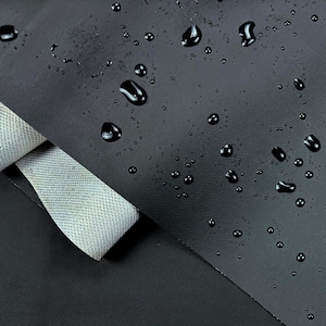 May include: Close-up of a black, textured material with water droplets scattered across the surface. A gray, woven ribbon is partially visible, rolled over the edge of the material. The image highlights the texture and water resistance of the fabric.