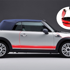 May include: A silver Mini Cooper convertible with a red and white stripe along the side. The car has a black fabric top and black wheels. The image shows a close-up of the red trim that can be used to protect the car's edges.