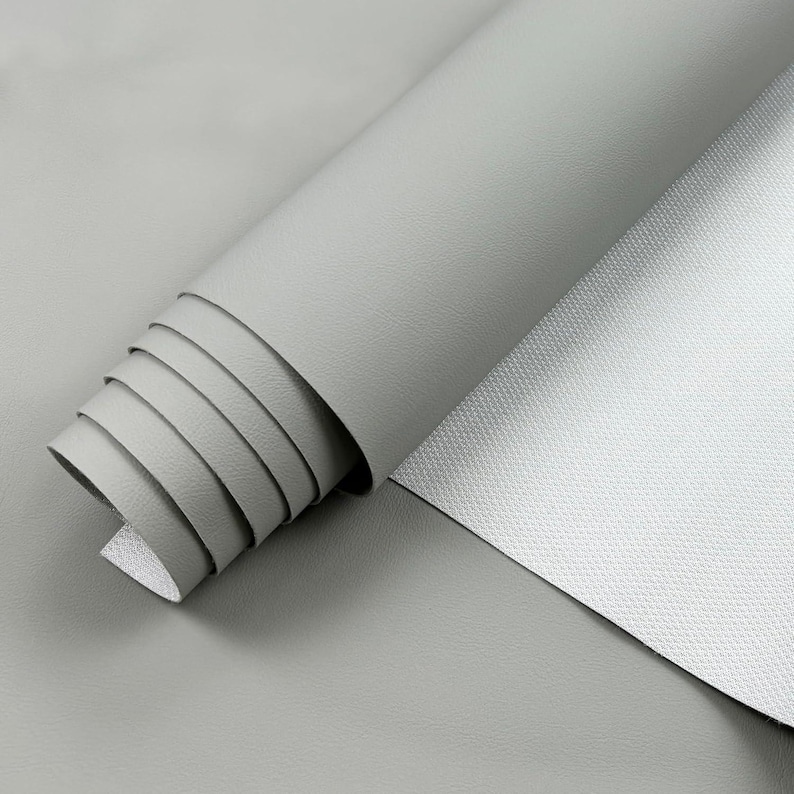 May include: A roll of light gray faux leather material, partially unrolled to reveal the textured surface. The material is smooth and appears to be new, suitable for crafting or upholstery projects.